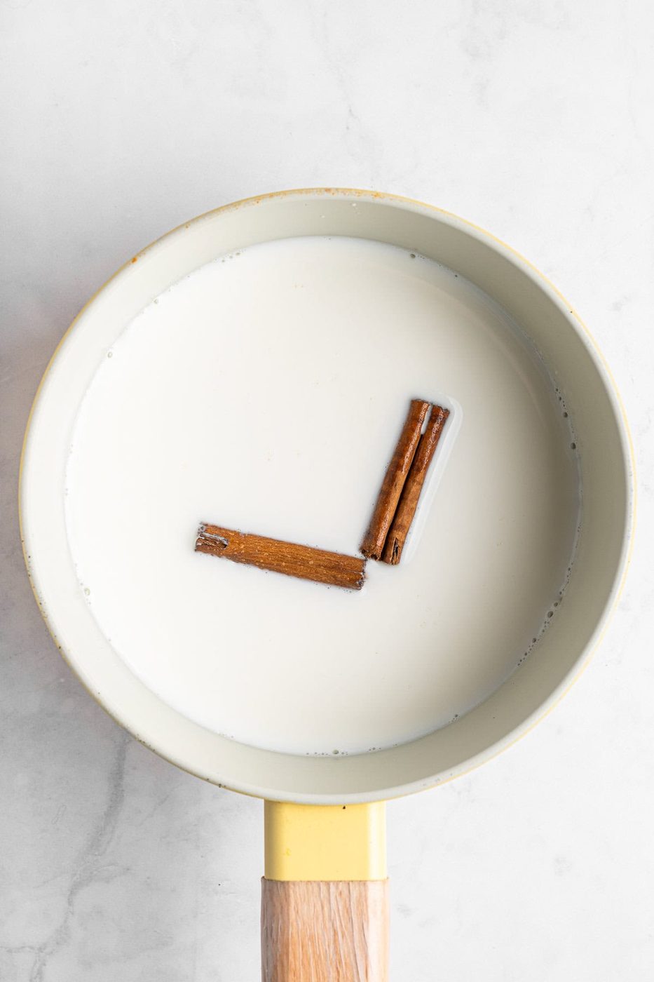 A saucepan filled with milk has two cinnamon sticks arranged to resemble a clock at 3 o'clock. The saucepan has a yellow exterior and a wooden handle, and rests on a light marble surface.