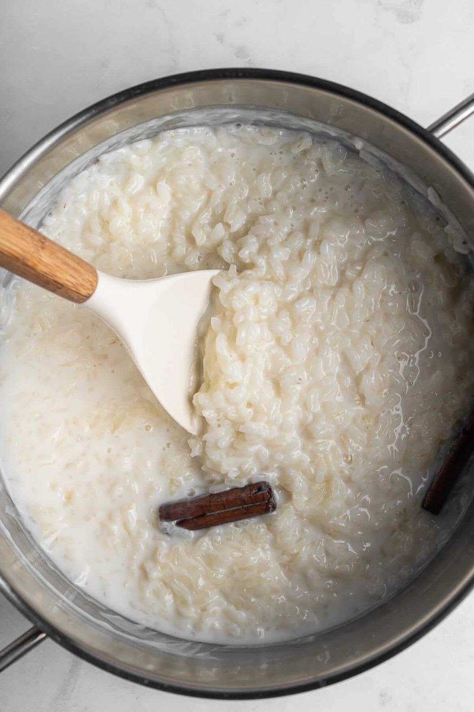 A pot of the creamy dessert. A cinnamon stick is partially submerged, and a white spatula with a wooden handle is resting in the pot. The background is a light marble surface.