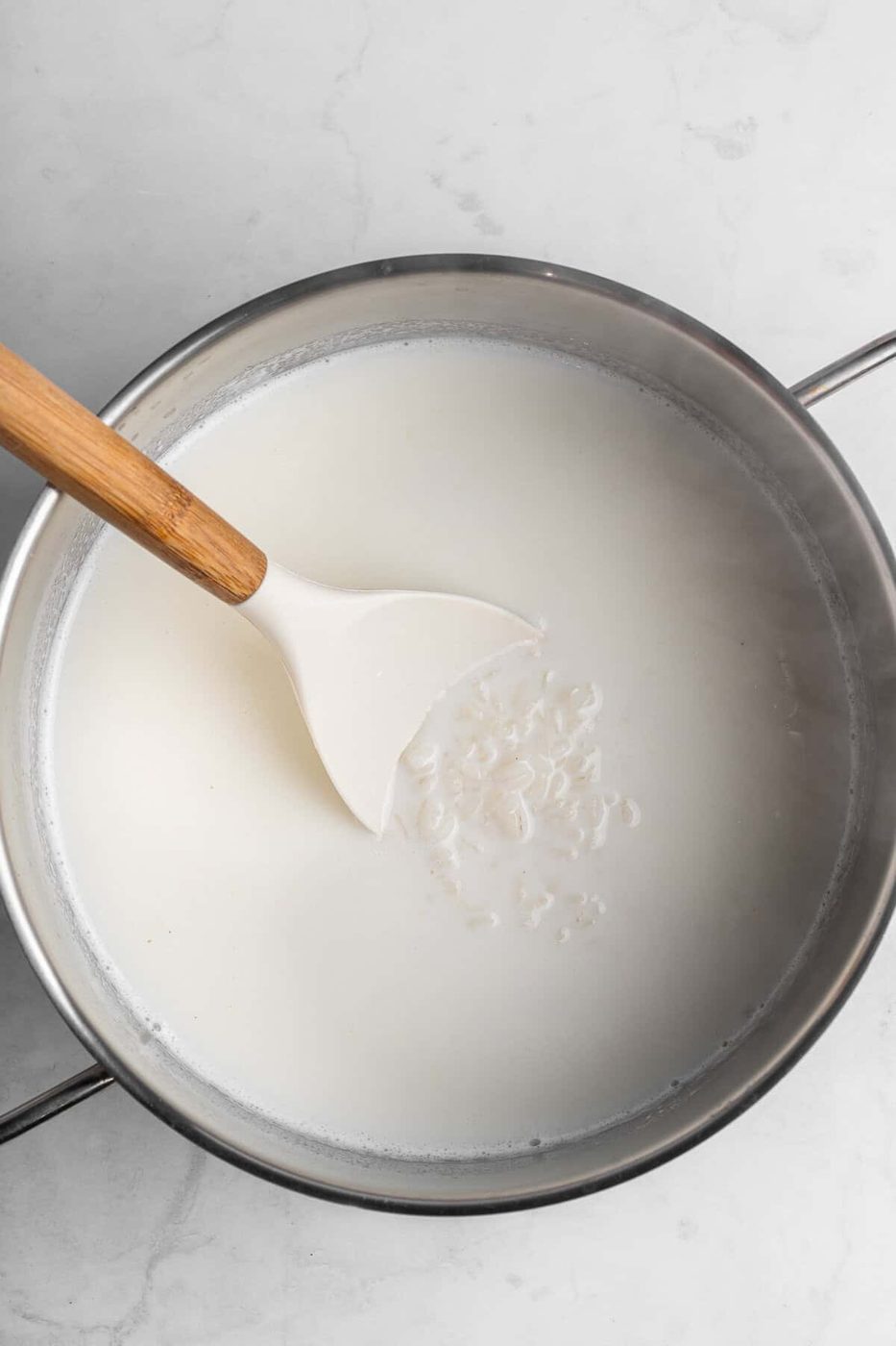 A pot filled with milk on a marble counter, with a white spatula stirring rice grains in the liquid.