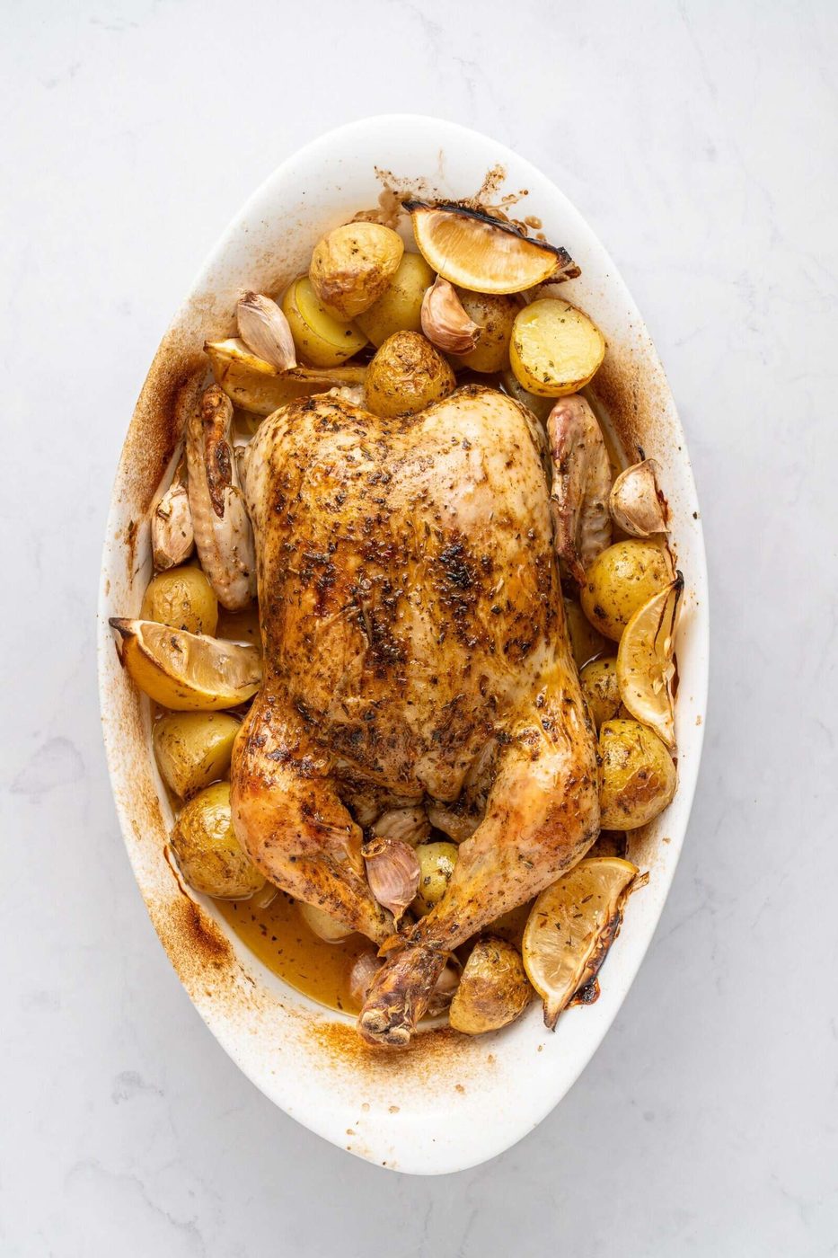Roasted whole chicken in an oval dish, surrounded by lemon wedges, garlic cloves, and golden potatoes. The dish is seasoned with visible herbs and has a golden brown, crispy texture. Placed on a light, marble surface.