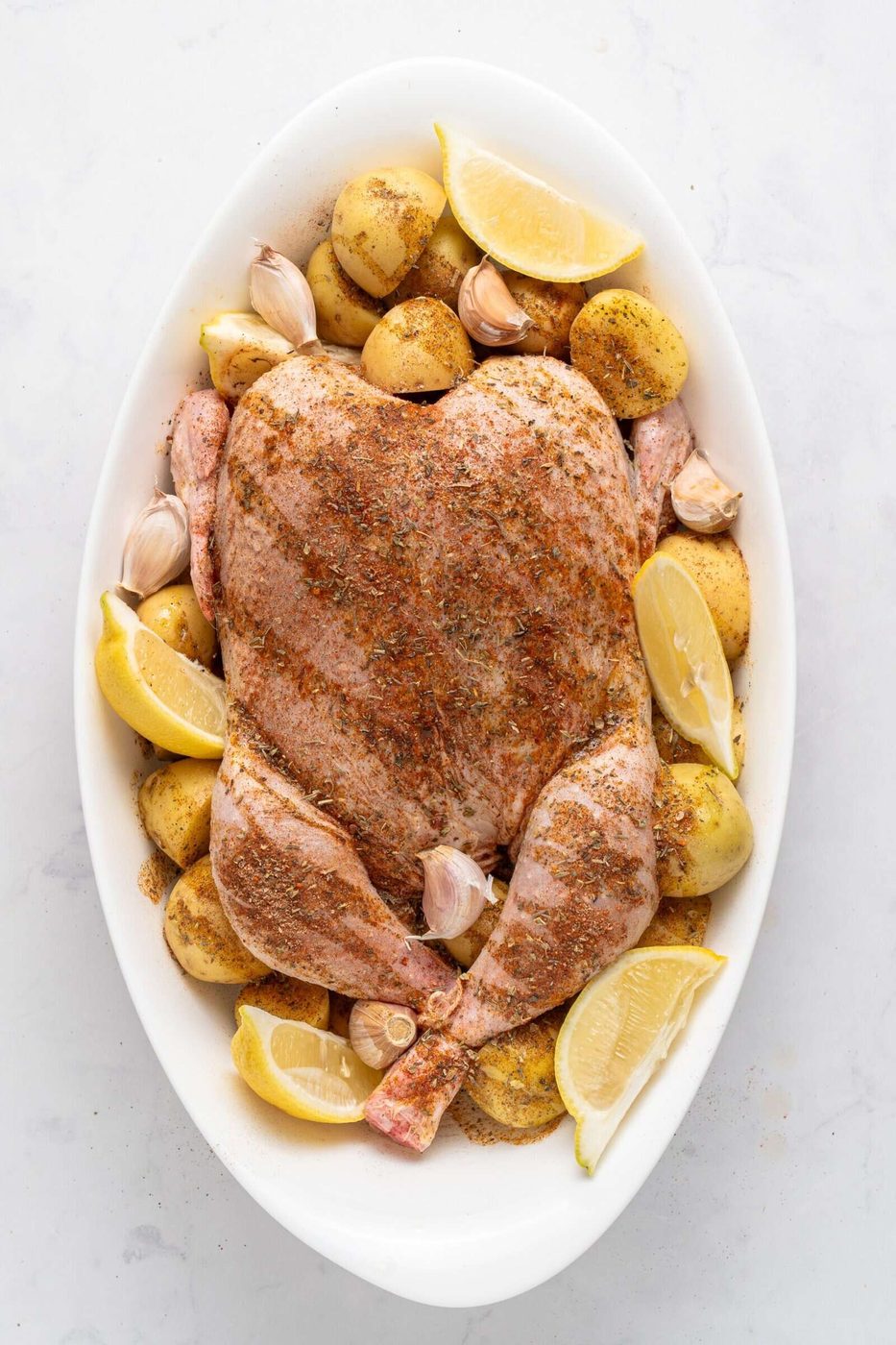A raw whole chicken, seasoned with spices, is placed in an oval dish. Surrounding it are halved small potatoes, garlic cloves, and lemon wedges, ready for roasting.