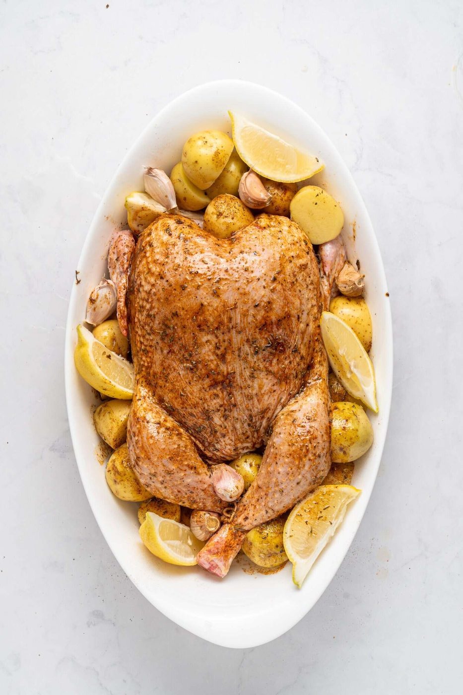 A roasted chicken seasoned with herbs is placed in an oval dish surrounded by roasted potatoes, lemon wedges, and garlic cloves on a light marble surface.