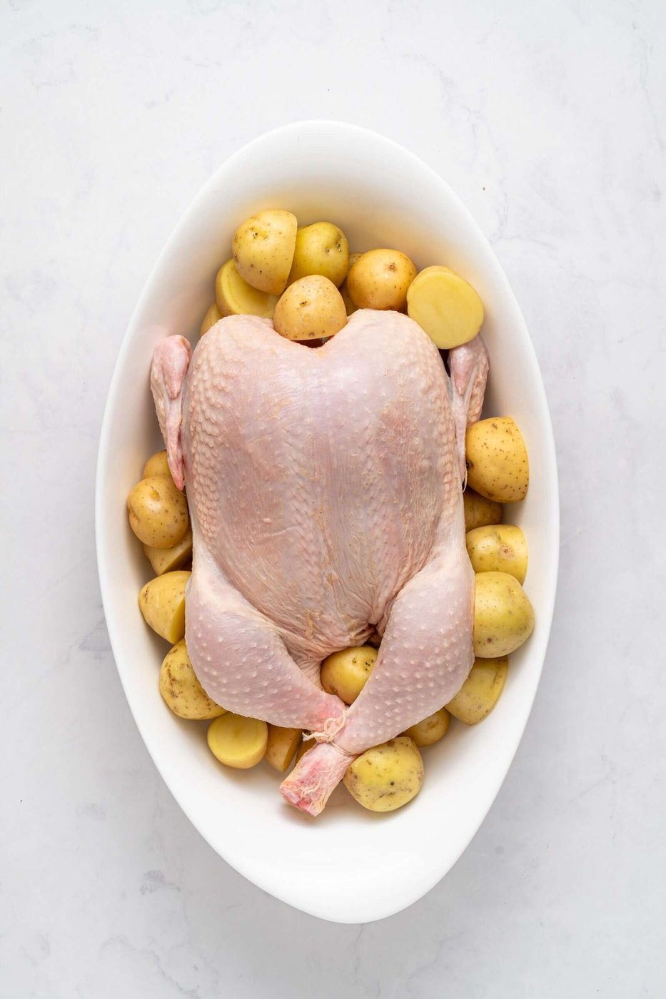 A raw whole chicken sits in an oval white dish, surrounded by halved baby potatoes. The dish is placed on a white marble surface.
