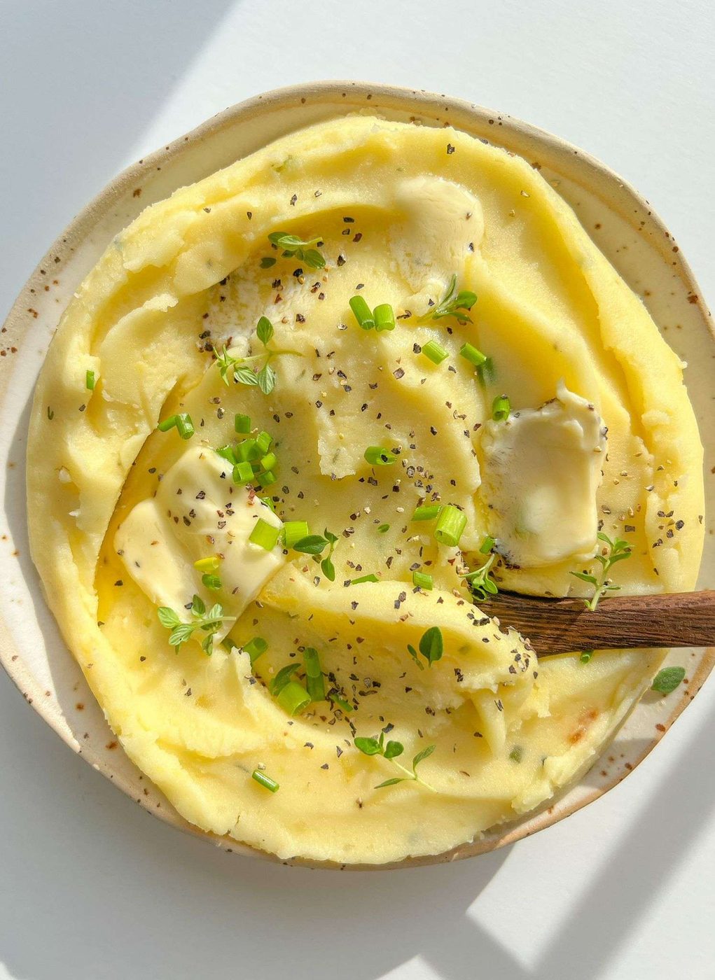 A plate of creamy Roasted Garlic Mashed Potatoes topped with butter, cracked black pepper, and fresh green herbs, with a wooden spoon resting in the dish.