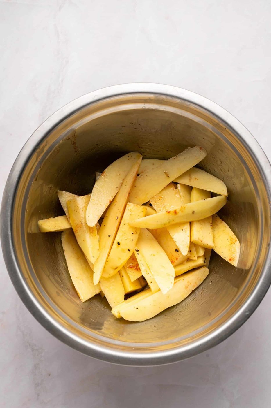 Raw potato wedges seasoned with spices in a stainless steel mixing bowl, placed on a light-colored countertop.
