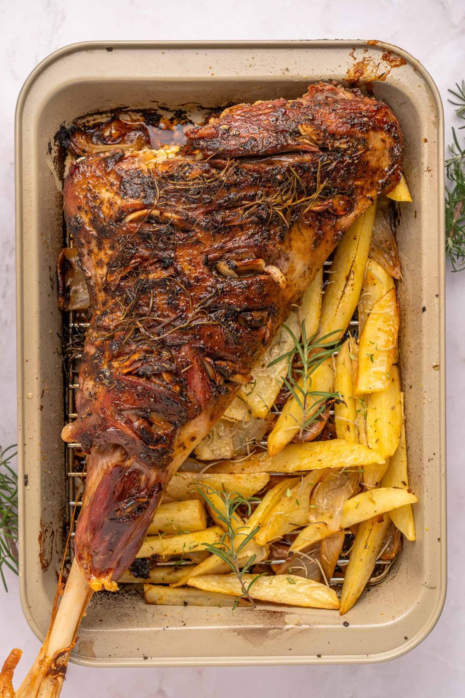 Roasted Leg of Lamb with herbs on a rack in a baking pan, surrounded by golden potato wedges and rosemary sprigs.