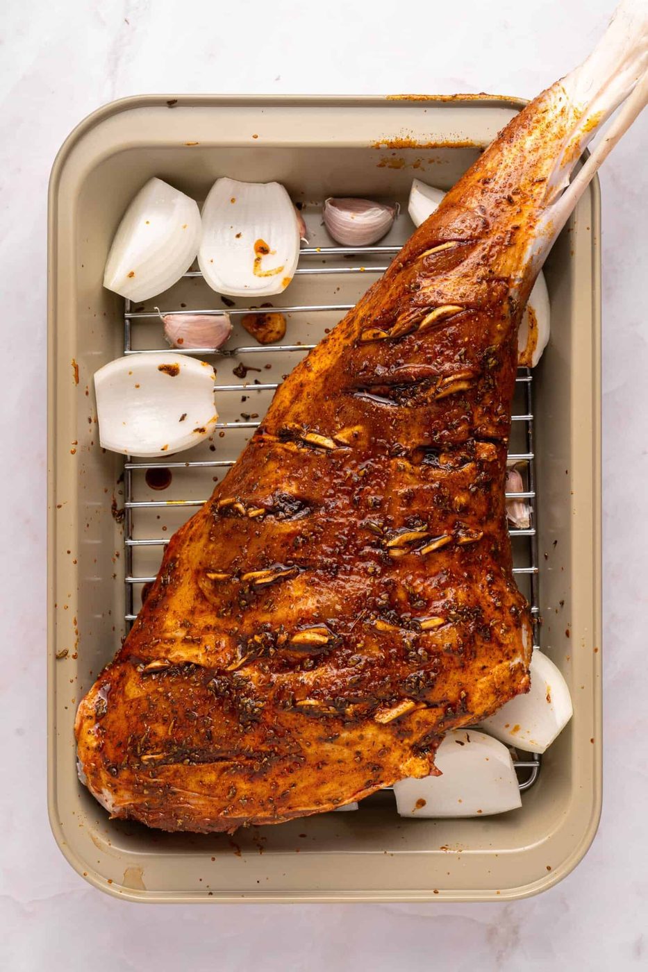 A seasoned, scored Roasted Leg of Lamb rests on a rack in a roasting pan, surrounded by halved onions and whole garlic cloves, ready to be cooked.