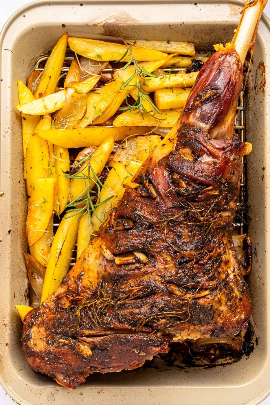 A roasted leg of lamb seasoned with herbs sits in a baking pan next to golden roasted potato wedges, garnished with sprigs of rosemary.