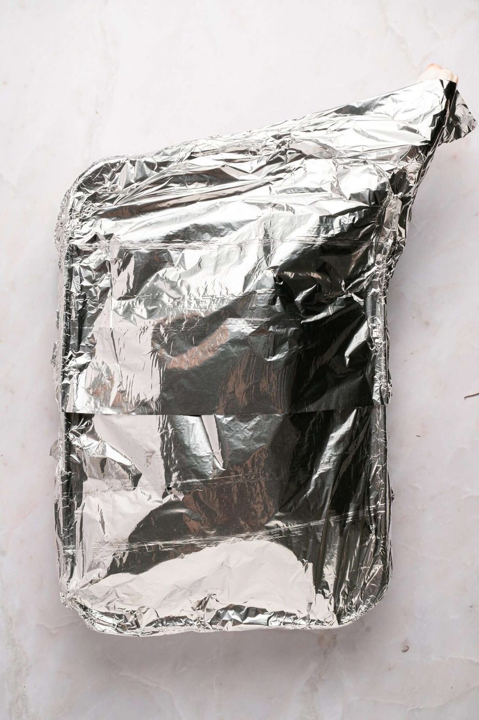A rectangular baking dish covered tightly with aluminum foil sits on a light-colored marble surface.