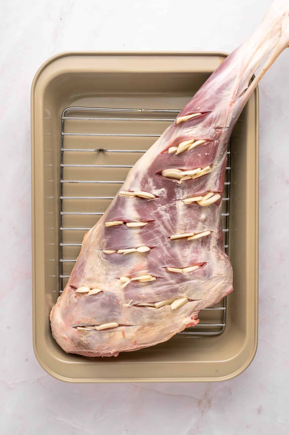 A raw leg of lamb with slits filled with garlic cloves, placed on a metal rack inside a beige roasting pan.