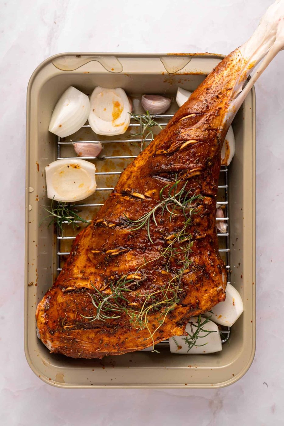 A seasoned and spiced leg of lamb sits on a rack in a roasting pan, surrounded by halved onions, garlic cloves, and sprigs of rosemary, ready to be cooked.