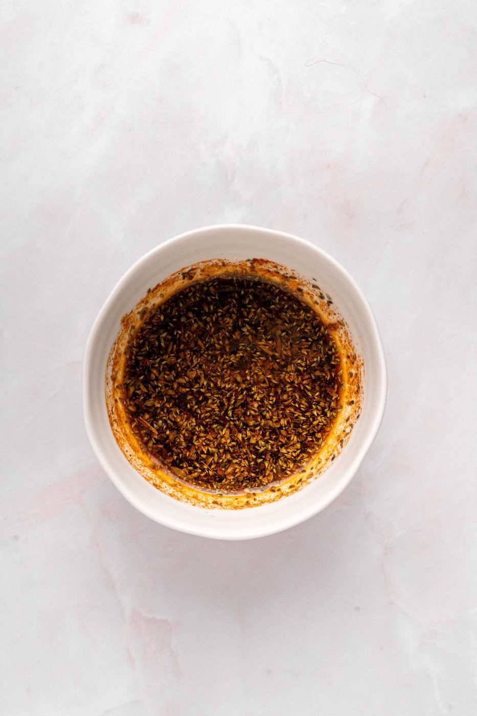 A white bowl filled with a dark, oily mixture of herbs and spices sits on a light marble surface.