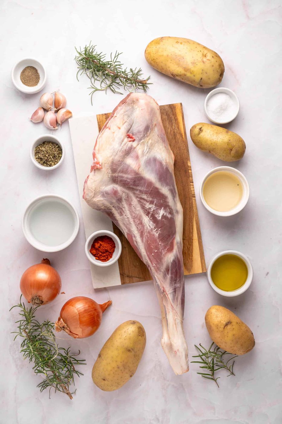 Raw lamb leg on a wooden board surrounded by potatoes, onions, garlic, rosemary, spices, salt, pepper, olive oil, and other seasonings in small bowls on a white background.