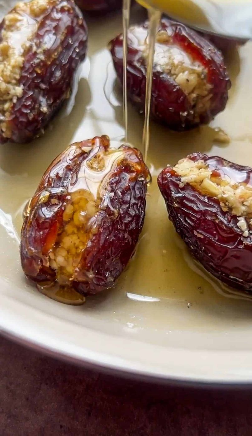 Close-up of a plate with stuffed dates drizzled with honey. The dates are filled with a nutty mixture, and a spoon is pouring honey over them, creating a glossy sheen. The background is softly blurred, focusing on the textured dates.