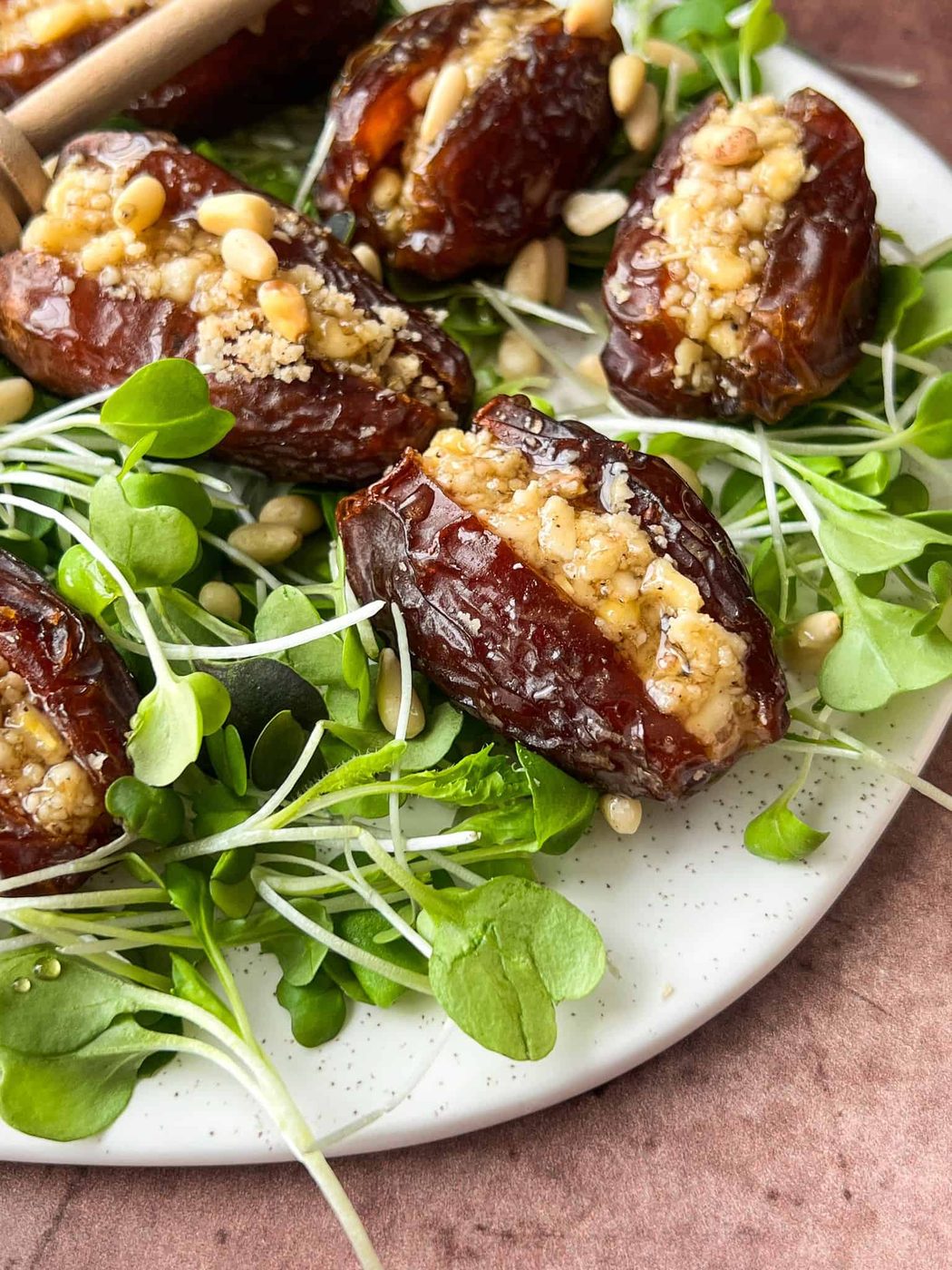 Dates stuffed with a nutty mixture are arranged on a plate with fresh greens and sprinkled with pine nuts. The dish is served on a textured, light-colored plate with a wooden background.