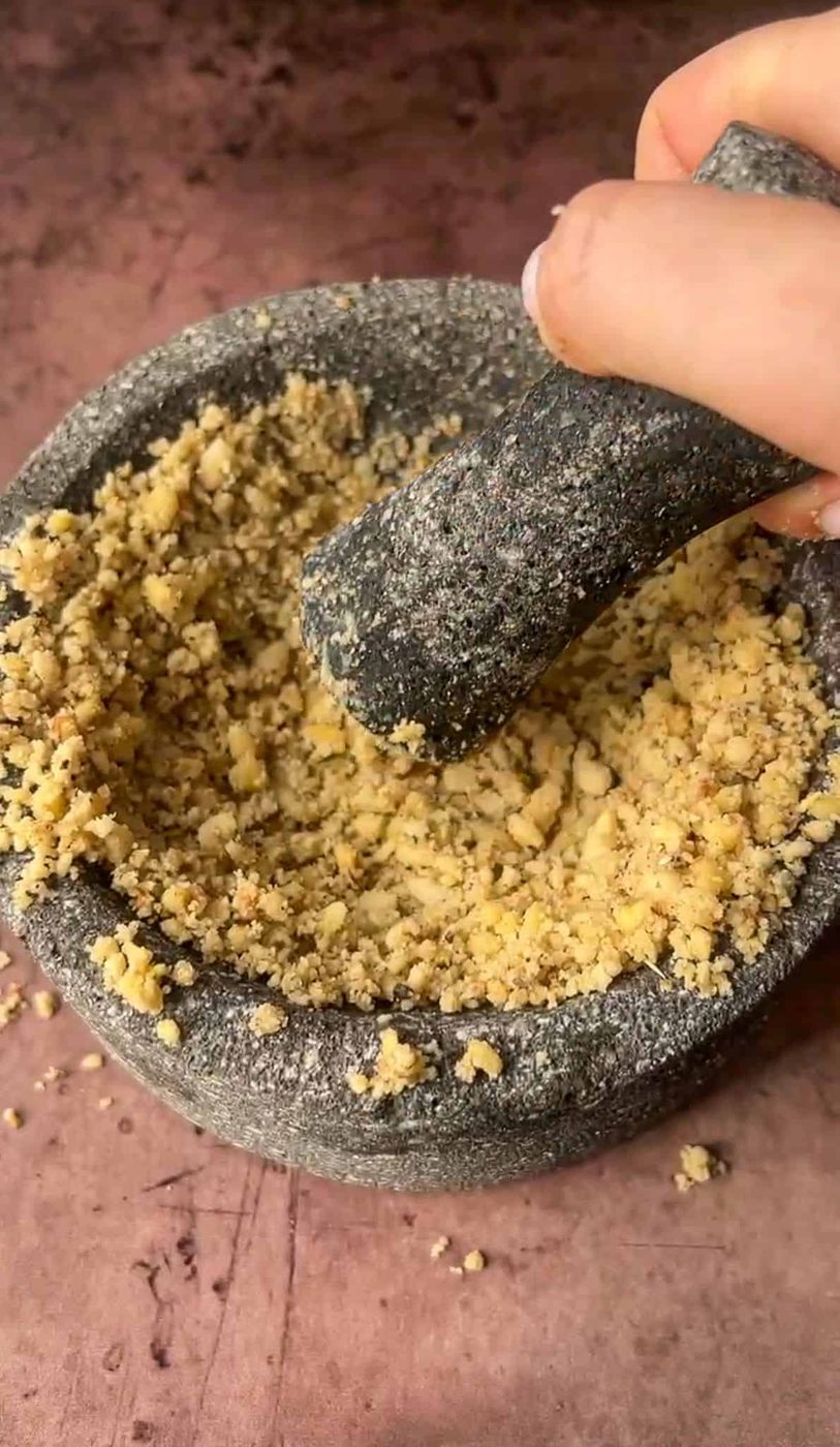 A hand using a pestle to grind nuts in a stone mortar. Crushed nuts fill the mortar, with some scattered on the table. The background is a brown textured surface.