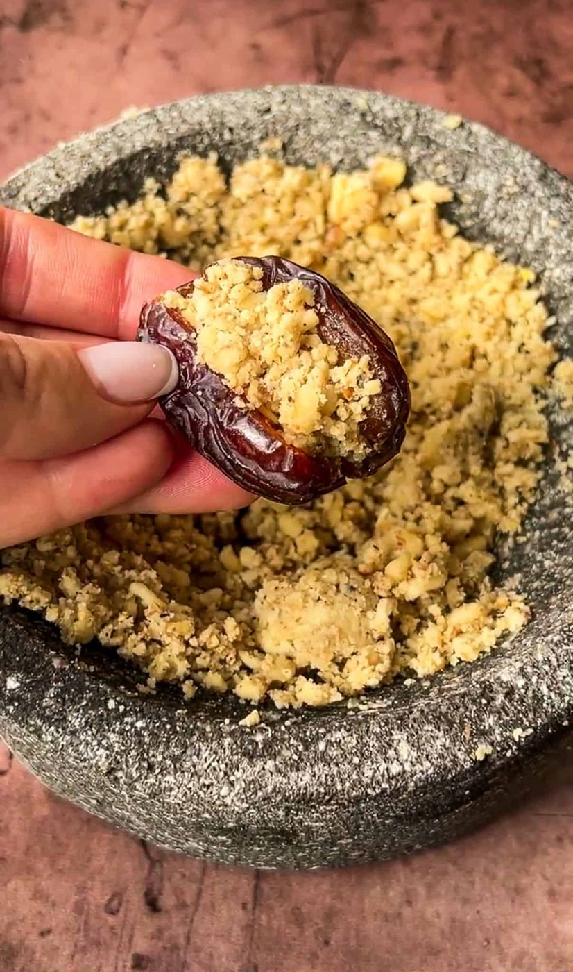 A hand holds an open date filled with crushed walnuts. Behind it, a stone mortar contains more crushed walnuts against a rustic brown surface.