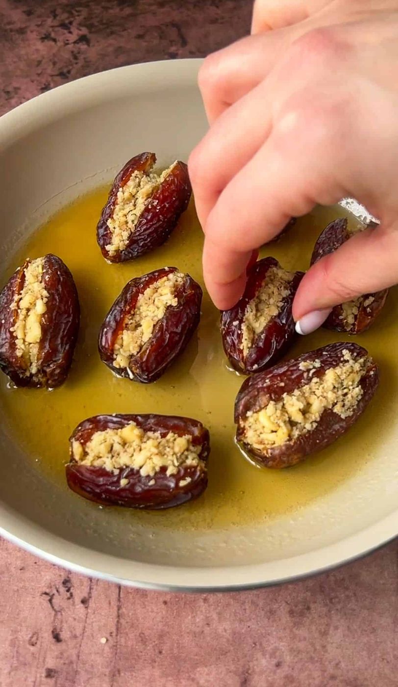 A hand is placing a stuffed date into a dish with several other dates. The dates are filled with a crumbly light brown mixture and are resting in a shallow layer of olive oil. The background surface is a textured brown.