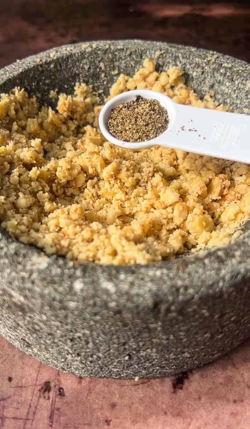 A stone bowl filled with crushed chickpeas. A white measuring spoon holds ground black pepper above the bowl, positioned to be added to the mixture. The background is a blurred, warm-toned surface.