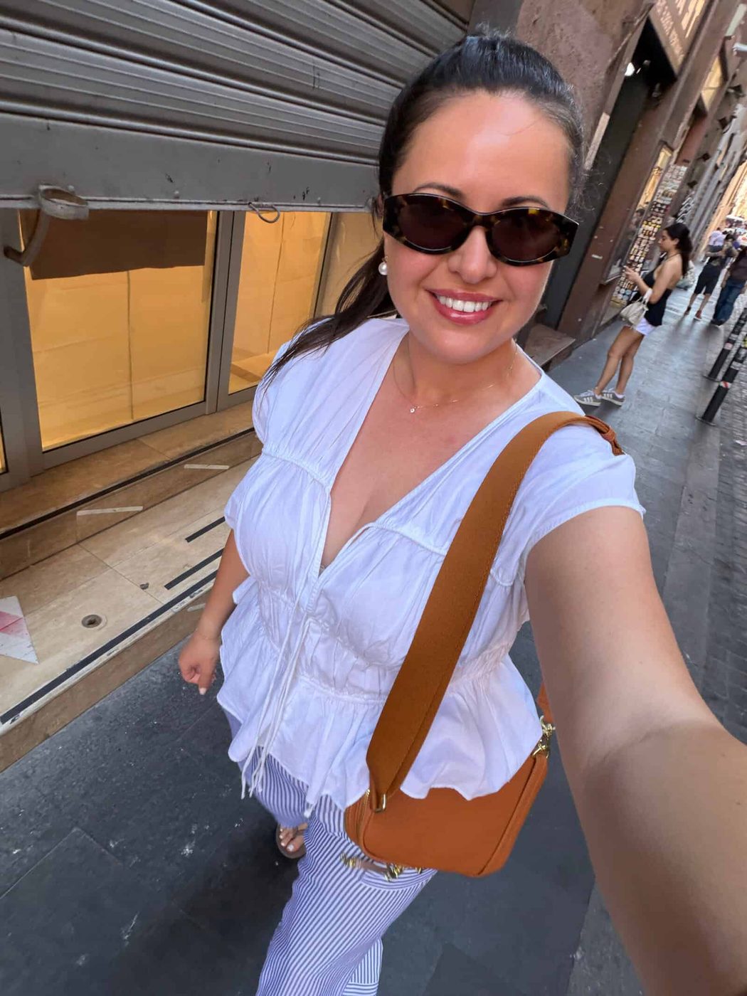 A woman wearing sunglasses, a white blouse, and striped pants takes a selfie while walking down a city street. She is smiling and carrying a brown shoulder bag. Shops and people are visible in the background.