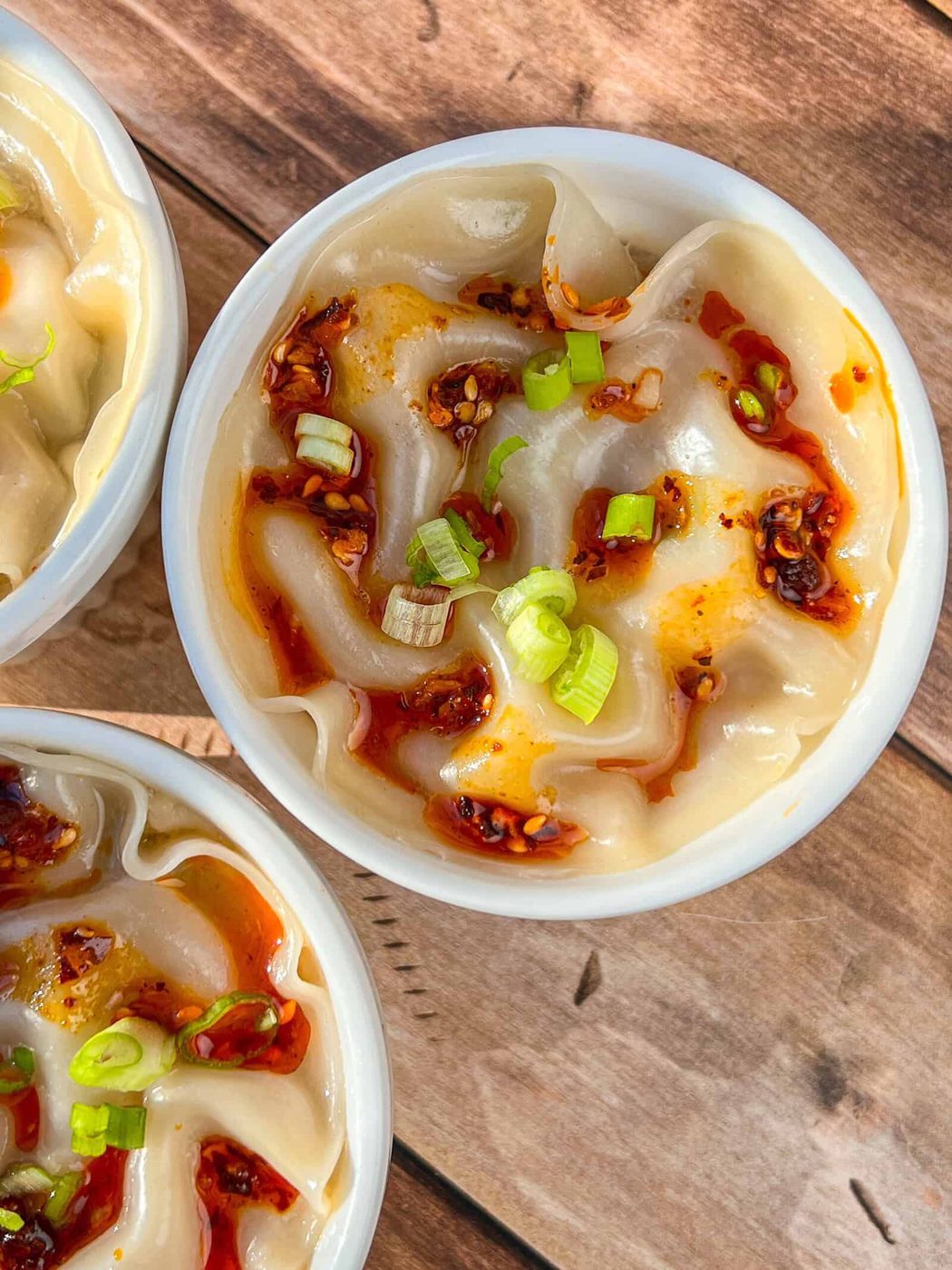A bowl of dumplings topped with chili oil and chopped green onions, placed on a wooden surface.