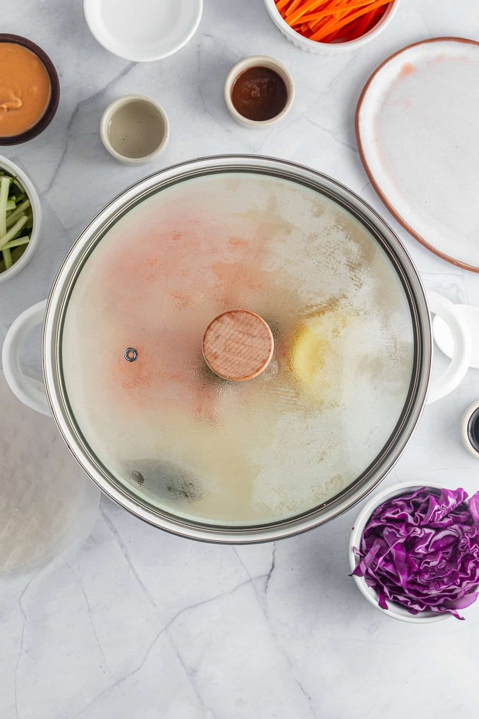 A pot with a glass lid is steaming on a marble countertop. Surrounding the pot are bowls of various ingredients, including sliced carrots, cucumbers, red cabbage, and small saucers of sauces, ready for cooking preparation.