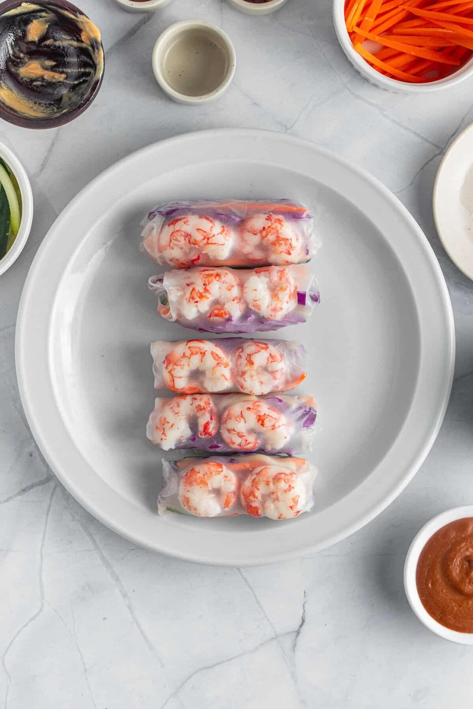 Four spring rolls are neatly arranged on a white plate. Surrounding the plate are small bowls containing various ingredients and sauces, including sliced vegetables and dipping sauces. The background is a marble surface.