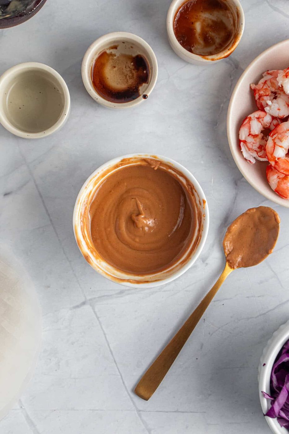 A top-down view of a creamy peanut sauce in a white bowl, surrounded by small bowls containing shrimp, sauce, and other ingredients on a light-colored marble surface. A wooden spoon with sauce rests beside the bowl.