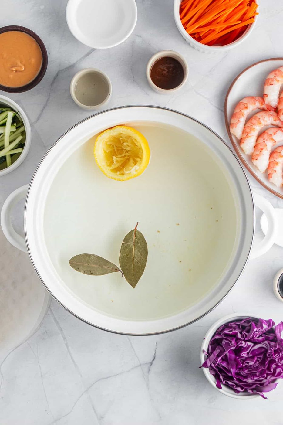 Ingredients for a dish are arranged around a white pot. The pot contains water with a lemon half and bay leaves. Surrounding the pot are shrimp, shredded purple cabbage, sliced carrots, and cucumber. Small bowls contain sauces and spices.