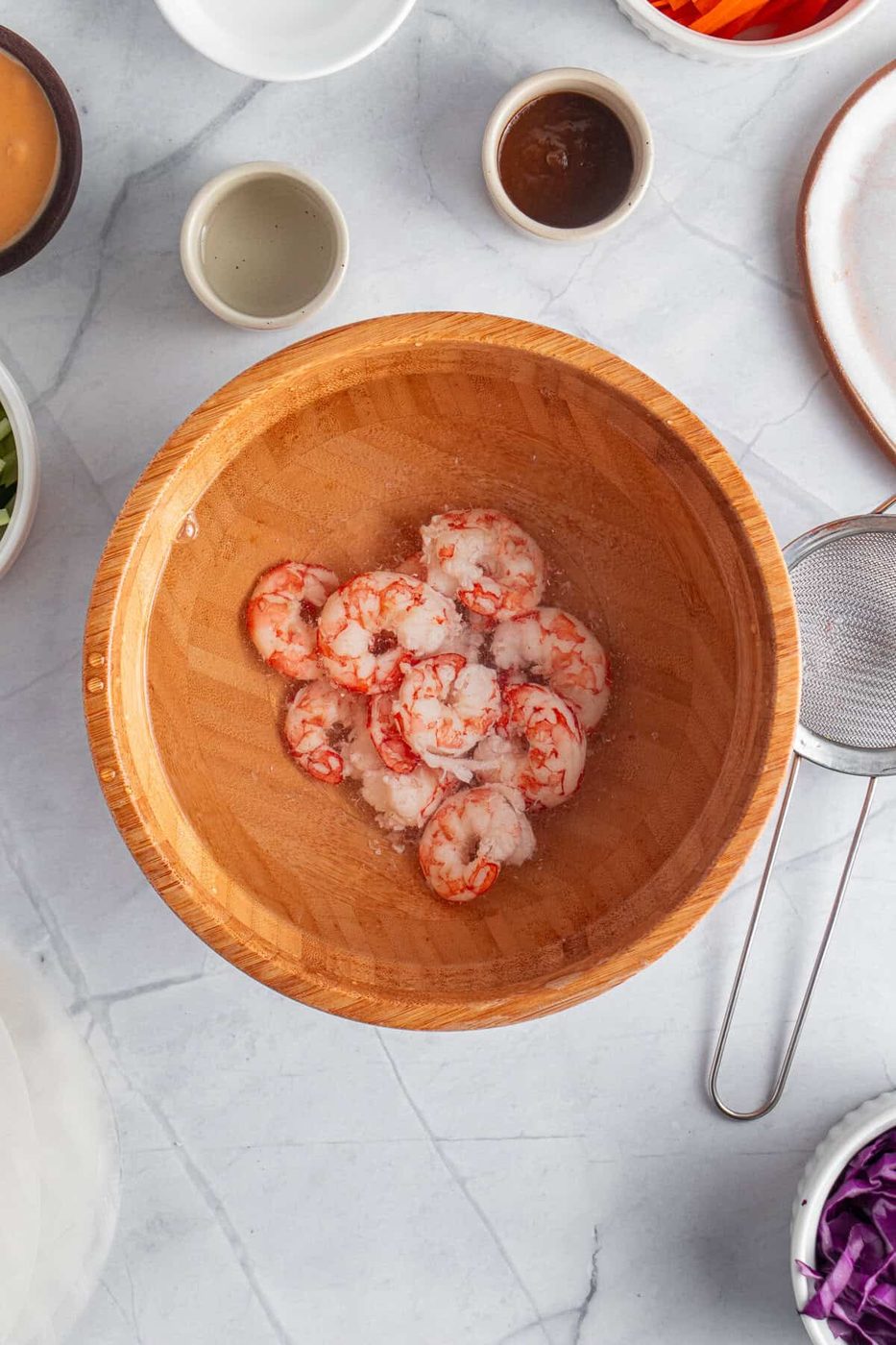A wooden bowl filled with shrimp sits on a marble countertop. Surrounding the bowl are small dishes with various ingredients and sauces, a strainer, and plates, suggesting preparations for a meal.