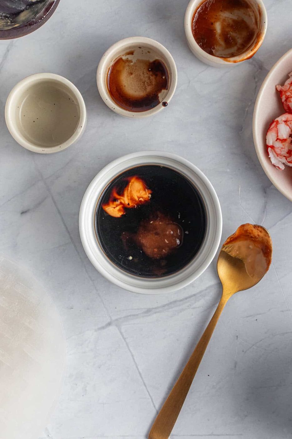 Various small bowls on a marble surface. One bowl contains a dark sauce with reddish garnish, and a golden spoon with sauce rests beside it. Other bowls hold remnants of ingredients and small bits of food, including shrimp.