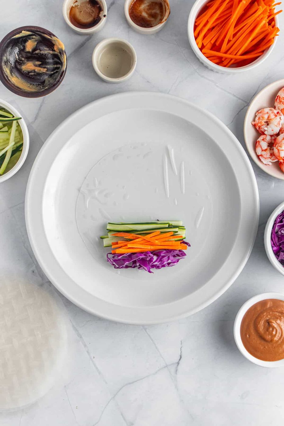 A plate with a rice paper sheet partially filled with julienned carrots, cucumbers, and purple cabbage, surrounded by bowls of shrimp, more vegetables, and various sauces, set on a light marble surface.