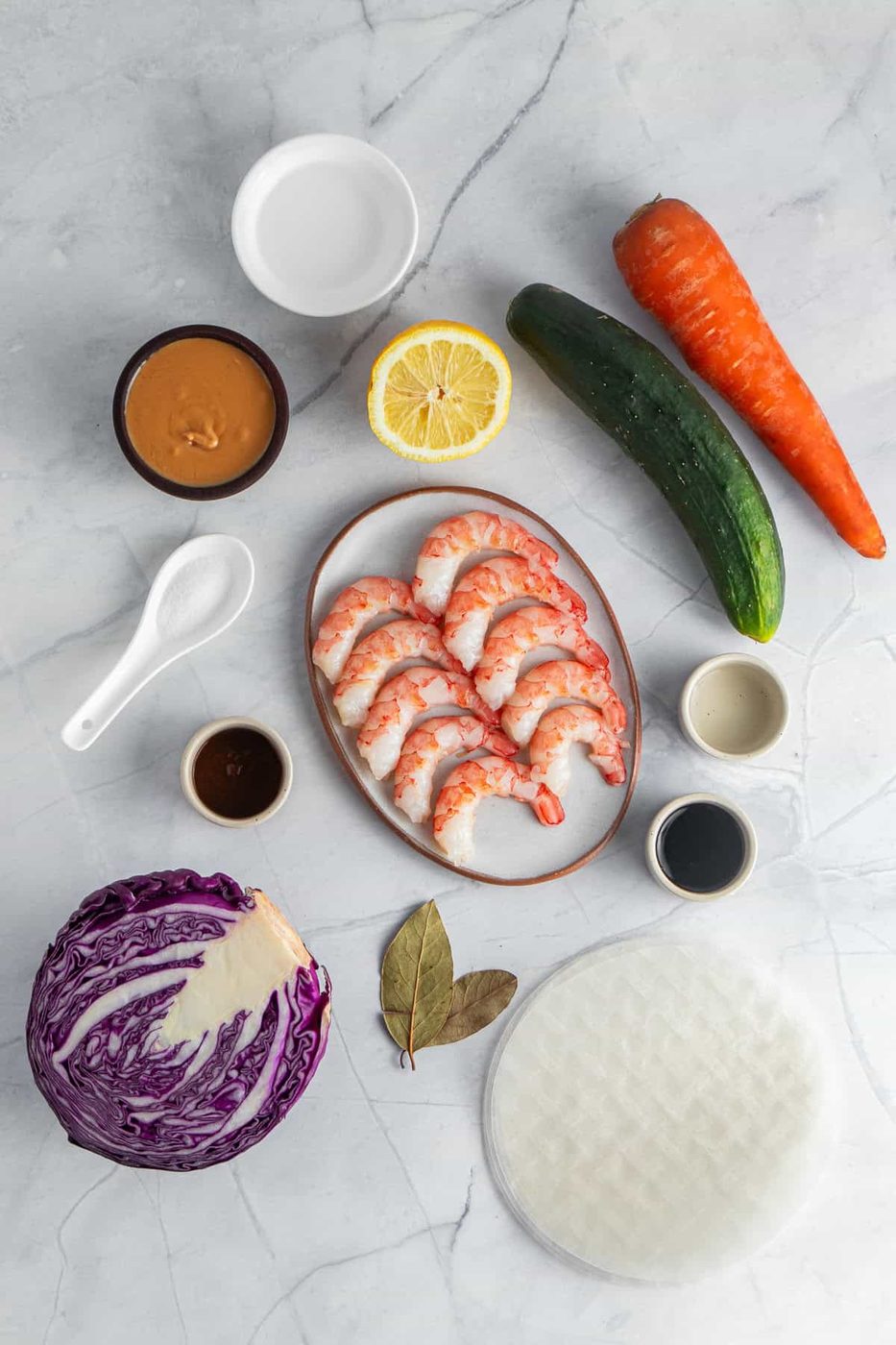 Ingredients for Vietnamese spring rolls on a marble surface: shrimp, carrot, cucumber, purple cabbage, lemon, rice paper, bay leaves, peanut sauce, soy sauce, hoisin sauce, and salt.