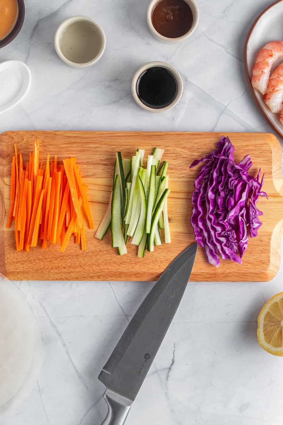 A wooden cutting board with sliced carrots, cucumbers, and shredded purple cabbage. A knife is placed nearby on the board. Surrounding the board are small bowls, shrimp, half a lemon, and a stack of rice paper.