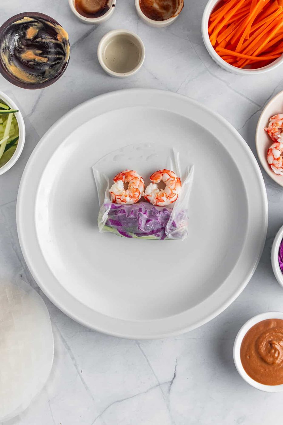 A partially assembled spring roll on a white plate, surrounded by bowls of ingredients like sliced carrots, cucumbers, shrimp, purple cabbage, and dipping sauces. A rice paper sheet is laid out next to the plate on a marble countertop.