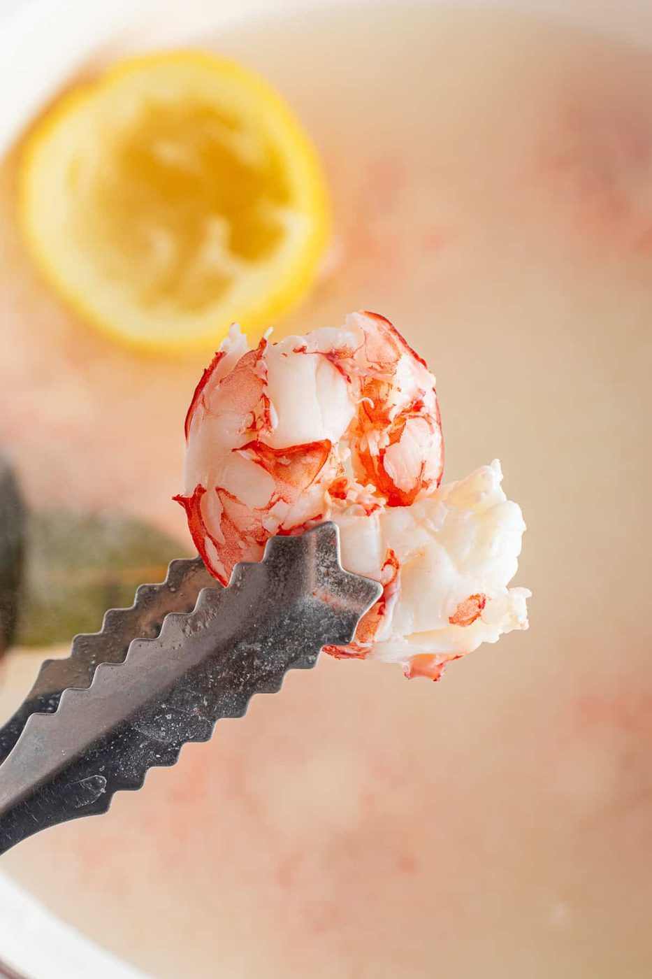 A close-up of a metal tong holding a piece of cooked lobster meat over a pot of boiling water. A half lemon is visible in the background. The lobster meat is white and red, showing its texture and freshness.