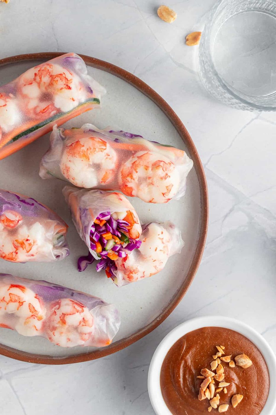 A plate of fresh spring rolls filled with shrimp, purple cabbage, and vegetables, partially unwrapped to reveal the colorful contents. Next to it, a bowl of peanut dipping sauce and a glass of water.
