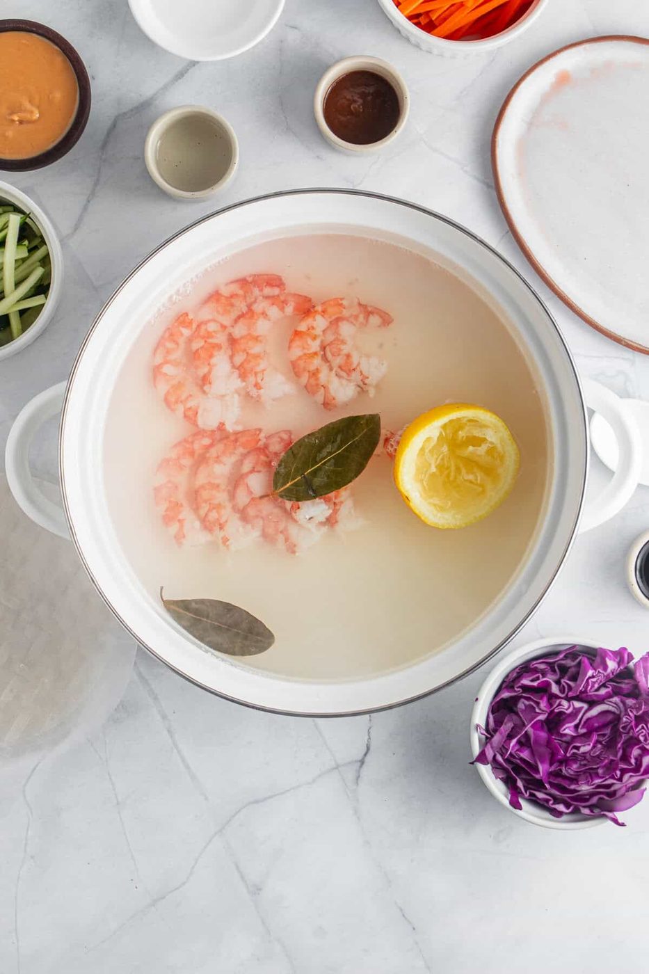 A pot filled with poaching water contains shrimp, a halved lemon, and bay leaves. Surrounding the pot are bowls with ingredients such as sliced vegetables, sauces, and spices on a marble countertop.