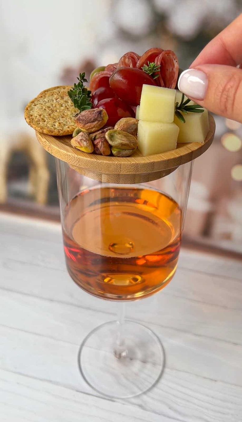 A glass of amber liquid with a wooden lid holding grapes, cheese cubes, crackers, pistachios, and a sprig of herbs. A hand is reaching to grab a cube of cheese with a toothpick.