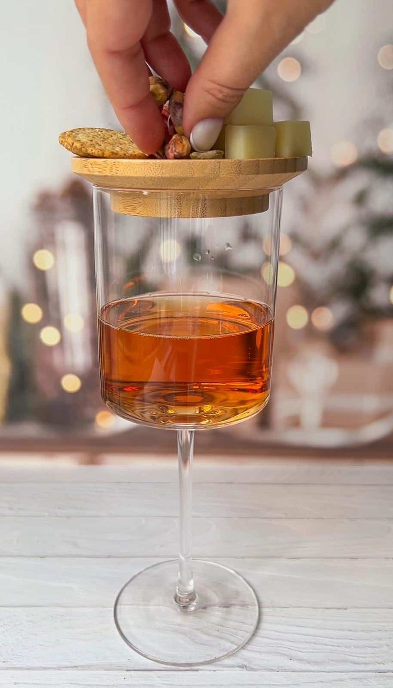 A hand places a wooden lid with assorted snacks, including grapes and crackers, on top of a glass filled with an amber liquid. The background is blurred with warm, soft lights.