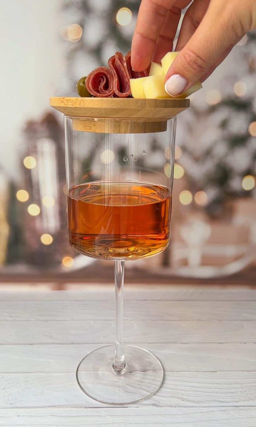 A hand places cheese cubes and a green olive on a wooden disc atop a wine glass filled with an amber beverage. The background is softly blurred with twinkling lights.