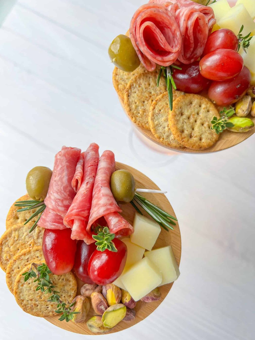 Two wooden platters with cured meats, crackers, green olives, cherry tomatoes, cheese cubes, pistachios, and garnished with rosemary, viewed from above on a light surface.
