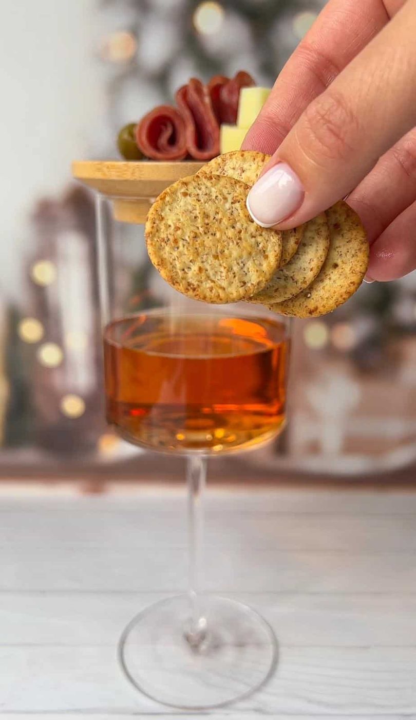 A hand holding three small round crackers over a glass of orange liquid. In the background, there's a wooden board topped with sliced meats like salami and some olives, slightly blurred. Holiday lights are softly glowing in the distance.