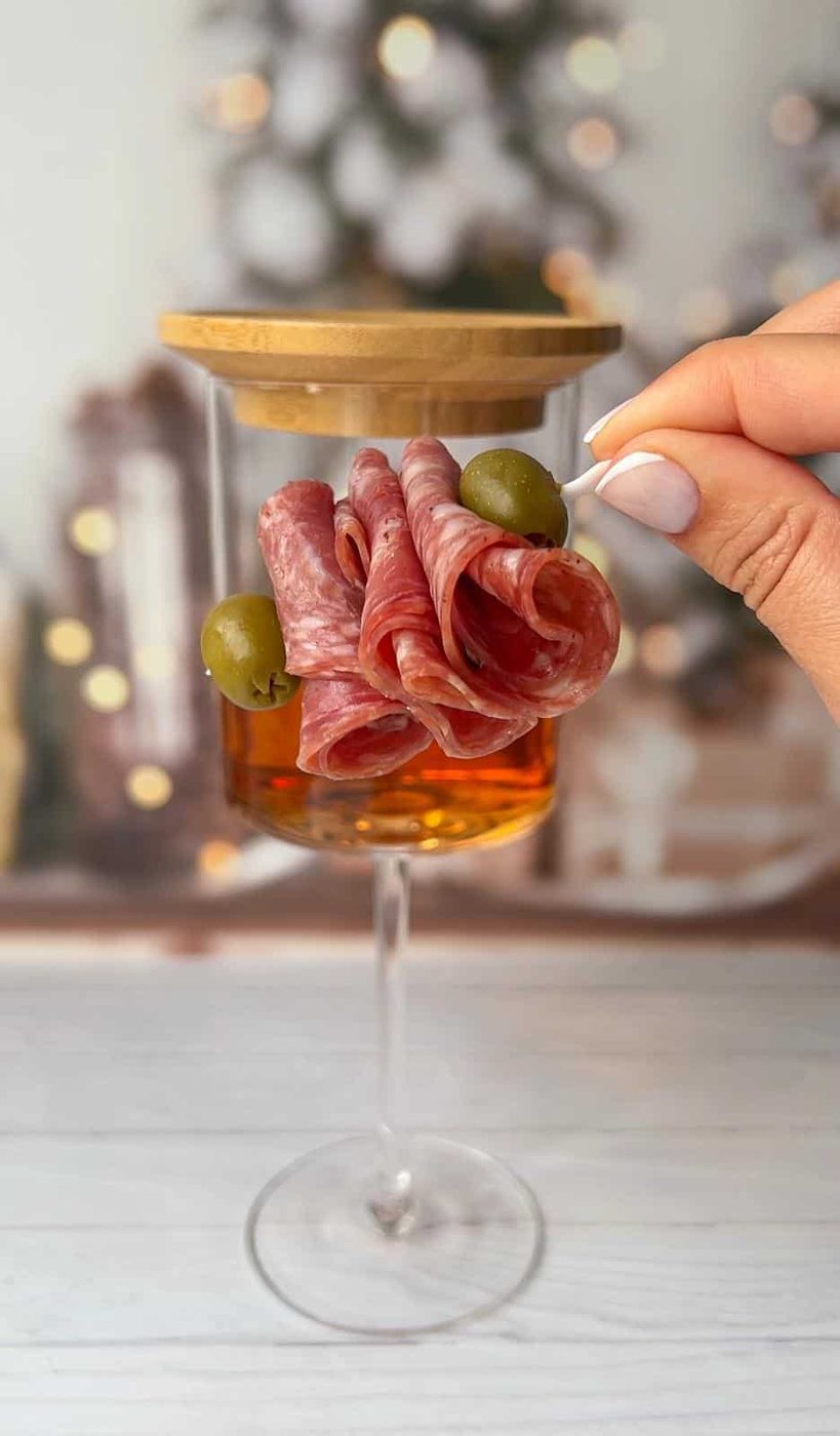 A hand holds a toothpick removing a green olive from a glass filled with rolled cured meats, which appears to be a creative charcuterie display. A wooden lid tops the glass. The background shows a blurred, warmly lit room.
