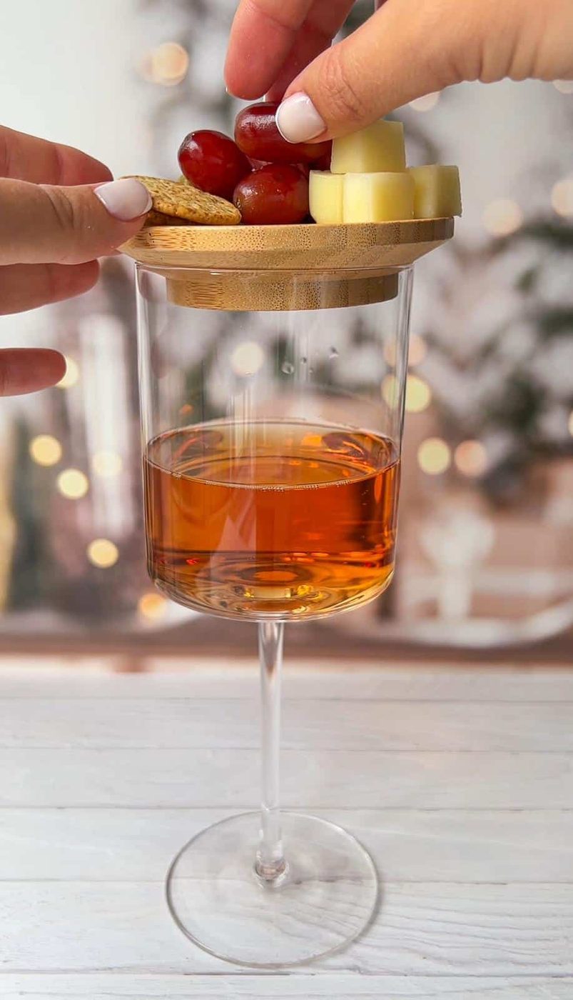 A glass of amber liquid with a wooden tray on top, holding grapes, cheese cubes, and crackers. A hand is adjusting the items on the tray. Blurred lights and decor are in the background.