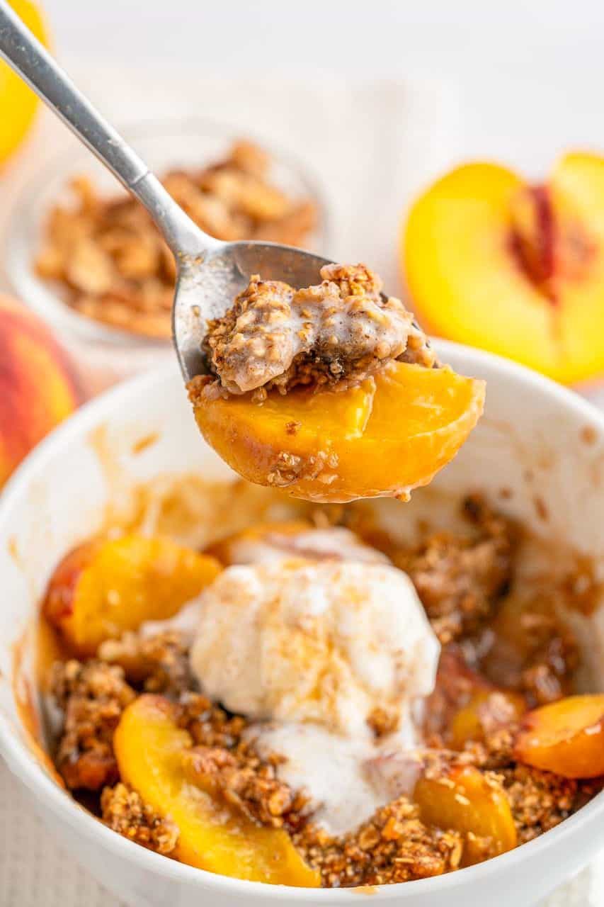 A photo of single serve peach crisp in a ramekin with baked peaches and ice cream on top.