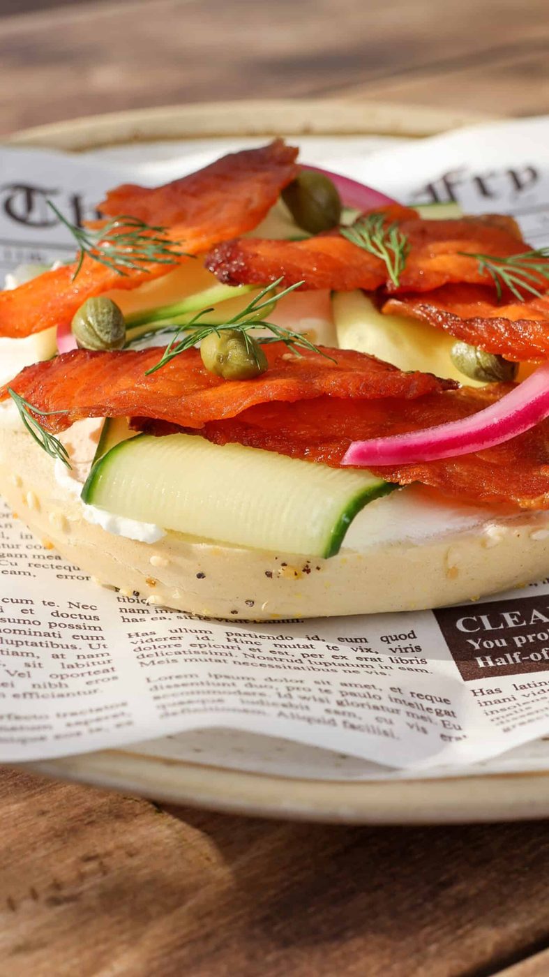 An open-faced bagel topped with cream cheese, smoked salmon jerky, capers, pickled red onions, fresh dill, and thin slices of zucchini, served on a plate lined with newspaper-print parchment.