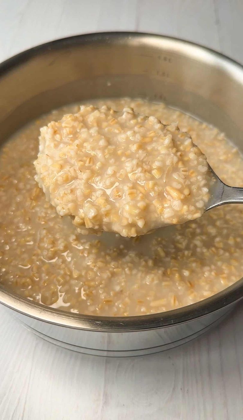 A close-up of a metal pan filled with cooked oatmeal. A spoon is lifting a portion of the oatmeal, showcasing its creamy texture. The background is a light-colored surface.