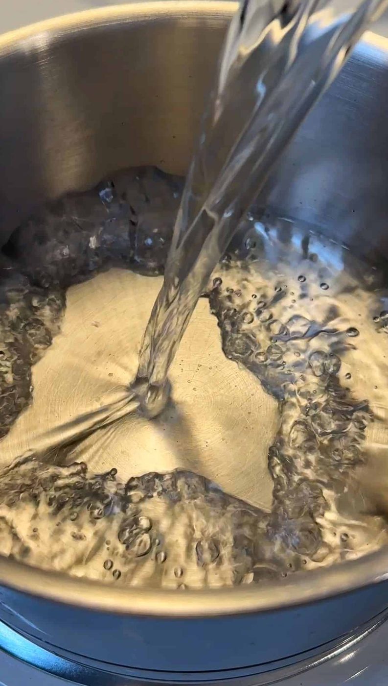 Water being poured into a stainless steel pot on a stove, with bubbles forming as it heats up.