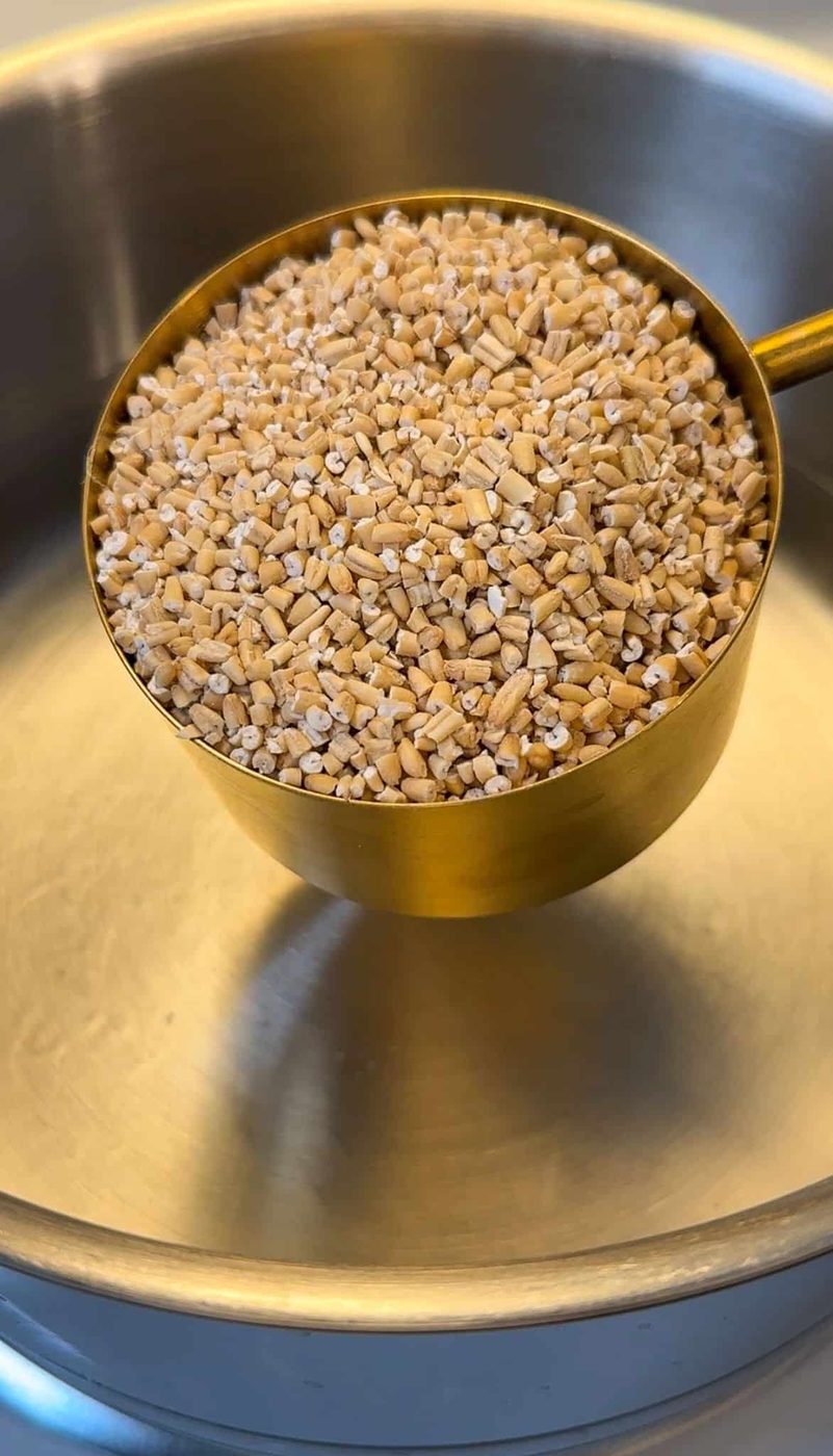 A measuring cup filled with steel-cut oats held above a shiny metal pot.